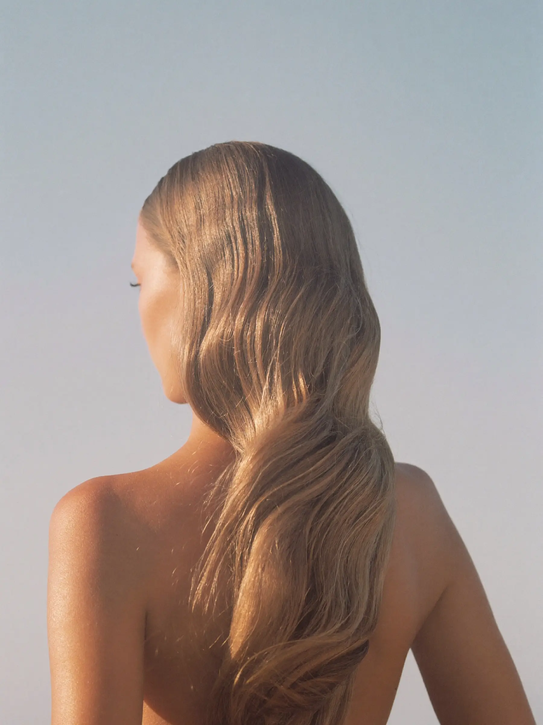 Mujer de espaldas con el cabello largo y rubio en ondas suaves, frente a un cielo claro
