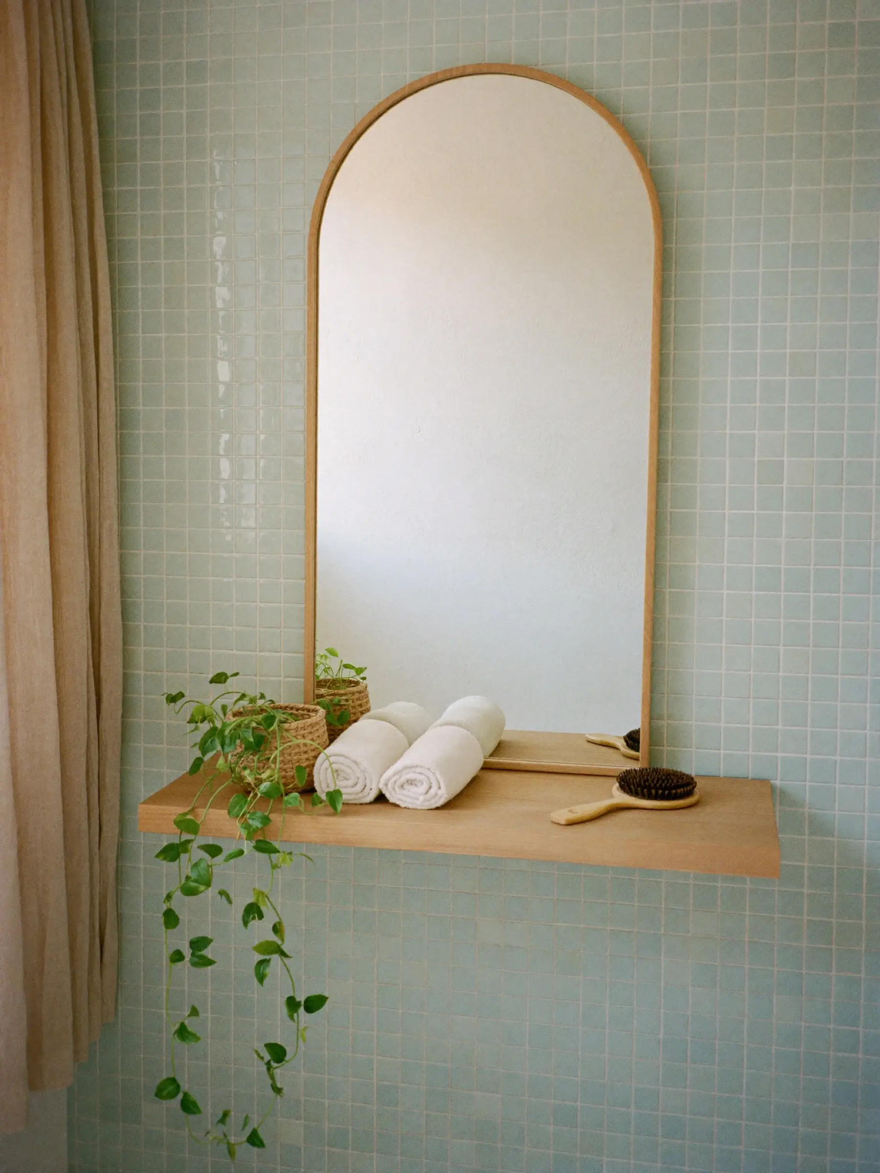 Espejo de arco con marco de madera clara sobre una pared de mosaico verde agua, con una repisa con planta, toallas blancas y un cepillo