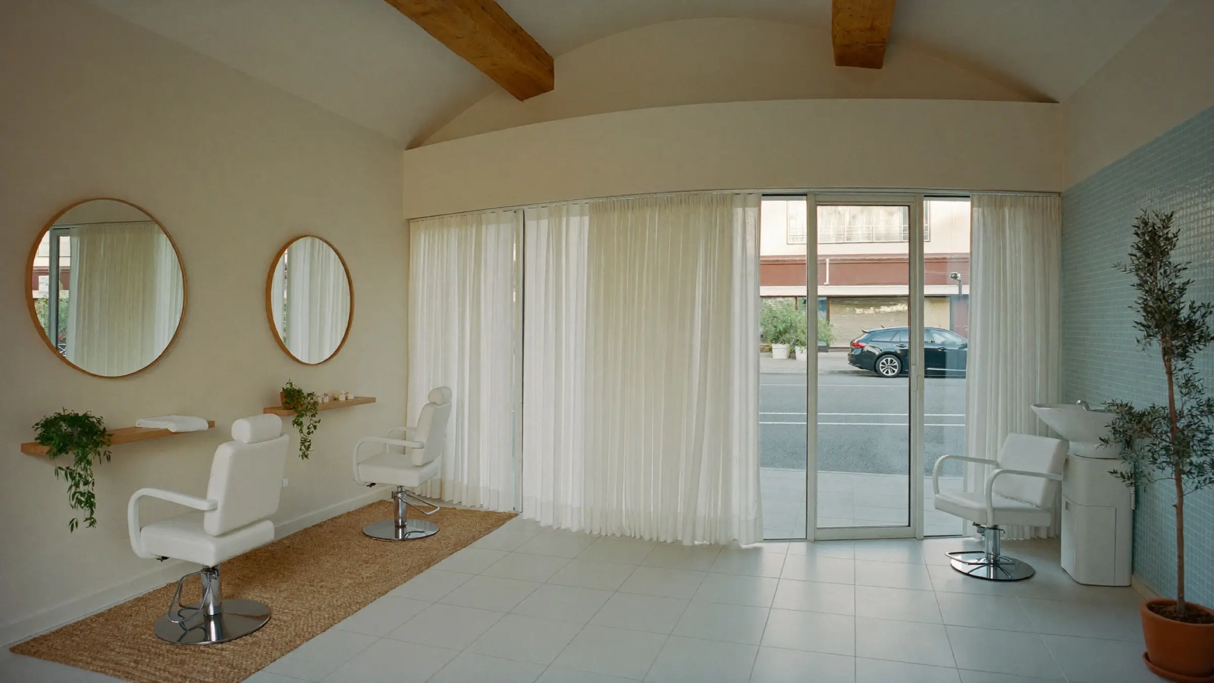 Salón luminoso con techo abovedado y vigas de madera clara, dos espejos redondos sobre repisas de madera con plantas, tres sillones blancos, cortinas blancas junto al ventanal y, a la derecha, un lavabo junto a un muro de mosaico azul verdoso con un olivo en maceta